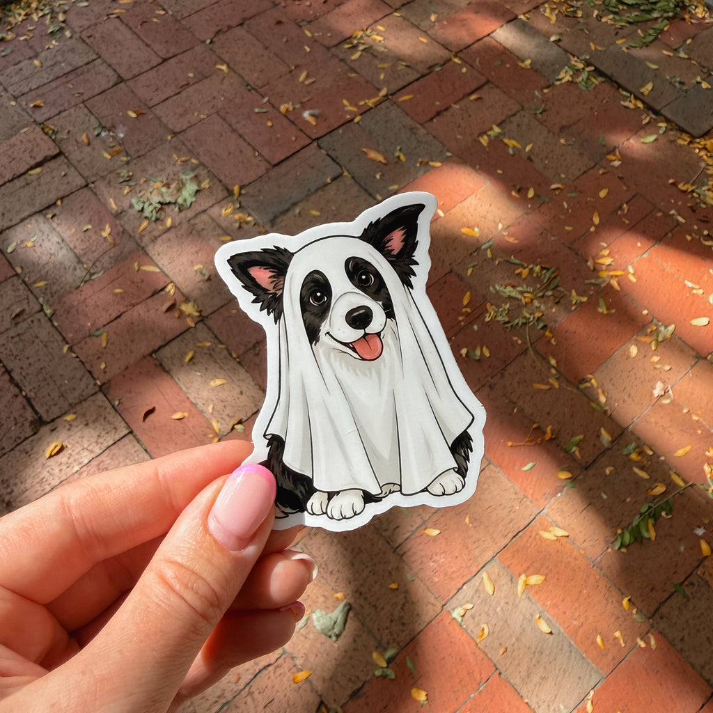 A hand holds a die-cut sticker of a black and white dog wearing a white ghost costume, set against a background of red brick pavement with scattered autumn leaves.