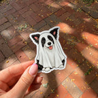 A hand holds a die-cut sticker of a black and white dog wearing a white ghost costume, set against a background of red brick pavement with scattered autumn leaves.