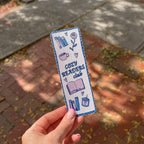 A hand with pink manicured nails holds a rectangular bookmark outdoors on a brick sidewalk. The bookmark has a white background with blue and purple illustrations of books, a cup, a flower, and the text 'COZY READERS club'.