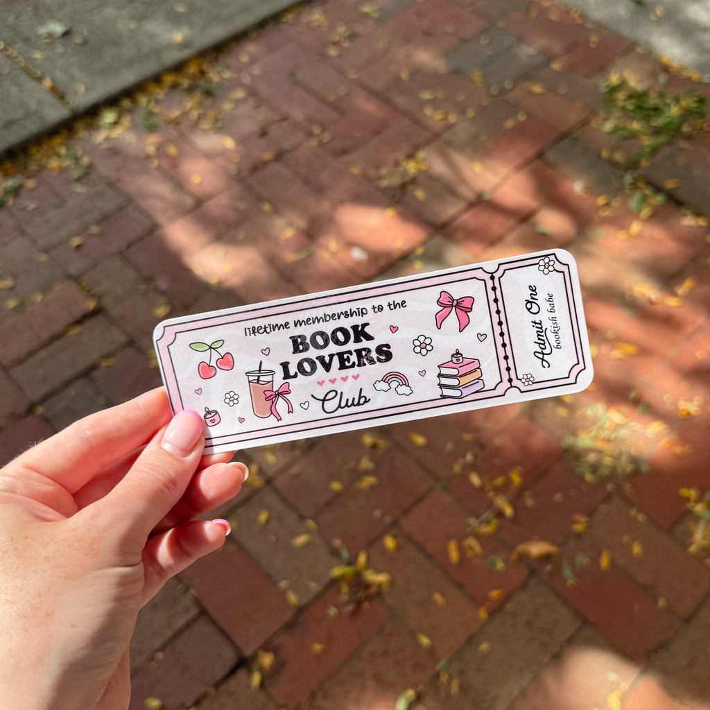 A hand holds a rectangular printed bookmark over a brick walkway. The card has a light pink background with black border lines and cute illustrations including cherries, a pink drink, a rainbow, a cake, and the text 'Welcome membership to the BOOK LOVERS Club' 