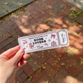 A hand holds a rectangular printed bookmark over a brick walkway. The card has a light pink background with black border lines and cute illustrations including cherries, a pink drink, a rainbow, a cake, and the text 'Welcome membership to the BOOK LOVERS Club' 