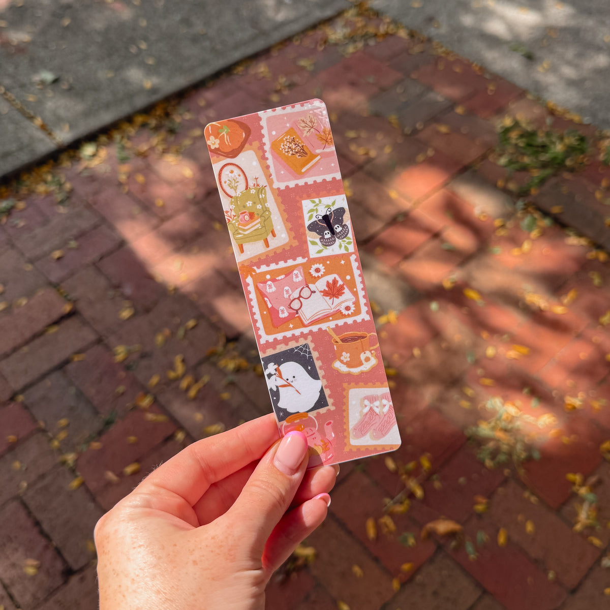 A hand holds a slim rectangular bookmark featuring a collage of autumn-themed illustrations, including a pumpkin, acorn, butterfly, book, and coffee cup, against an outdoor brick walkway background.
