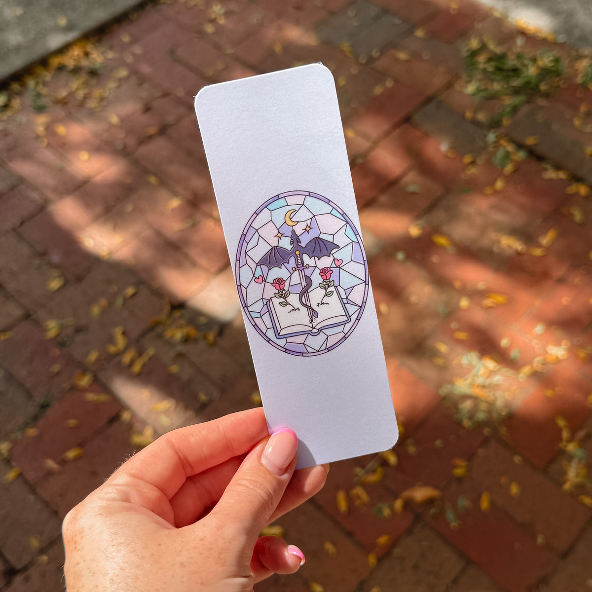 A hand holds a rectangular bookmark featuring a stained-glass-style illustration of a dragon, a crescent moon, and an open book with red roses. The background shows a blurred brick patio with scattered leaves in natural daylight.
