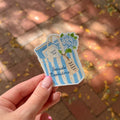 A hand with pink manicured nails holds a die-cut sticker shaped like a blue and white striped shopping bag. The sticker features the text 'Seaside Bookstore' and a blue hydrangea illustration, with a blurred brick patio in the background.