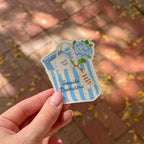 A hand with pink manicured nails holds a die-cut sticker shaped like a blue and white striped shopping bag. The sticker features the text 'Seaside Bookstore' and a blue hydrangea illustration, with a blurred brick patio in the background.