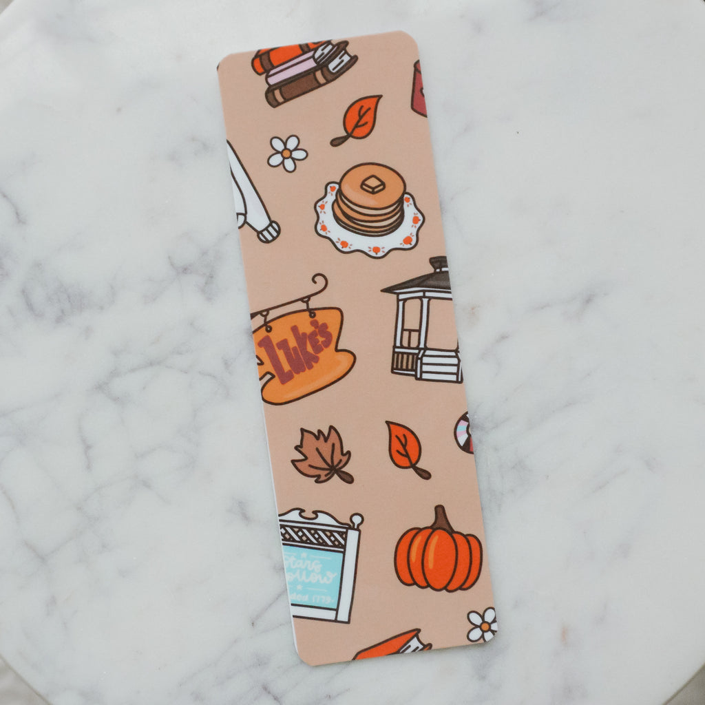 A rectangular metal bookmark with rounded corners featuring a light peach background and an autumn-themed illustration pattern, placed on a white marble surface.