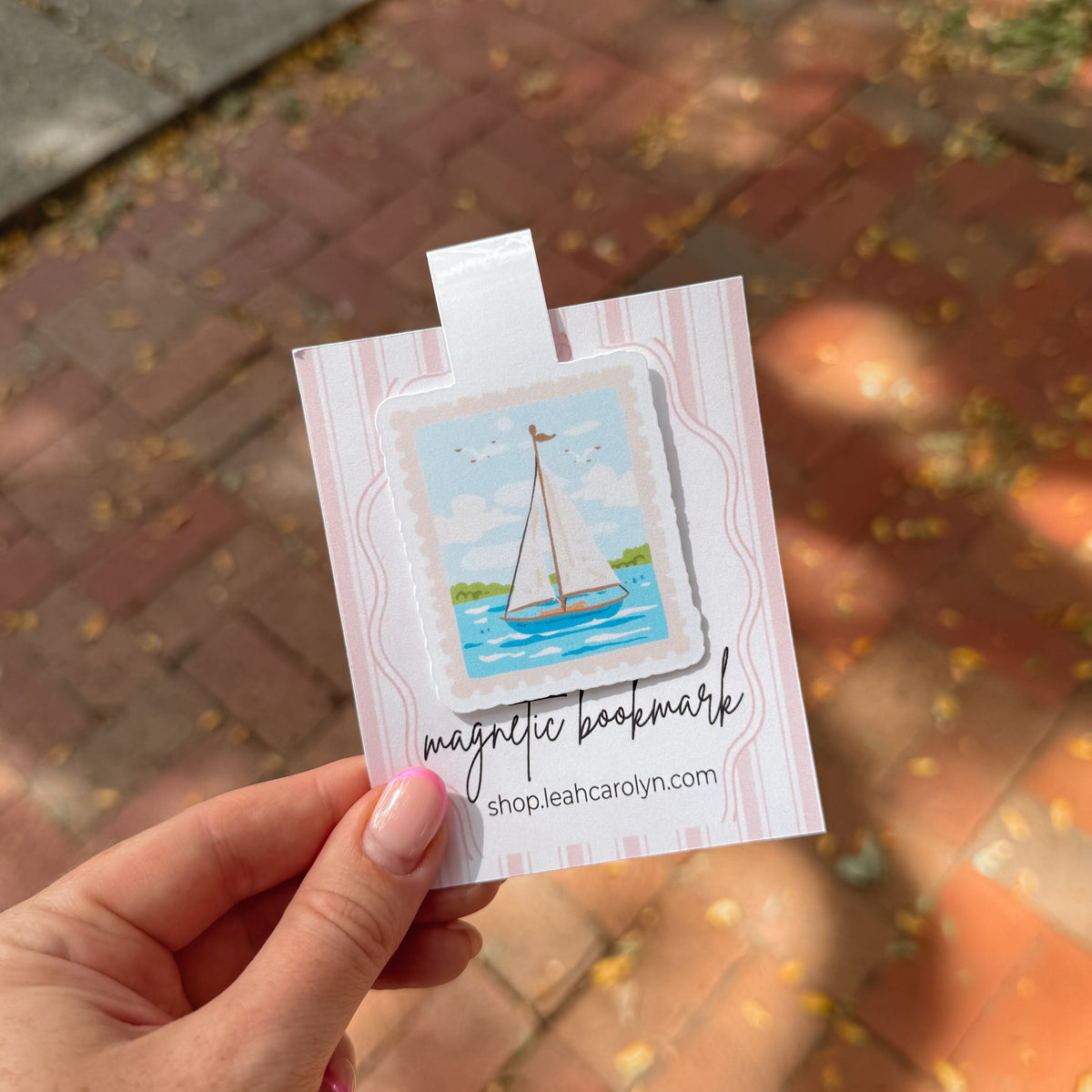 A hand holds a magnetic bookmark featuring a colorful illustration of a sailboat on blue water, attached to a pink and white striped backing card with the text 'magnetic bookmark' and 'shopleahcarolyn.com'.
