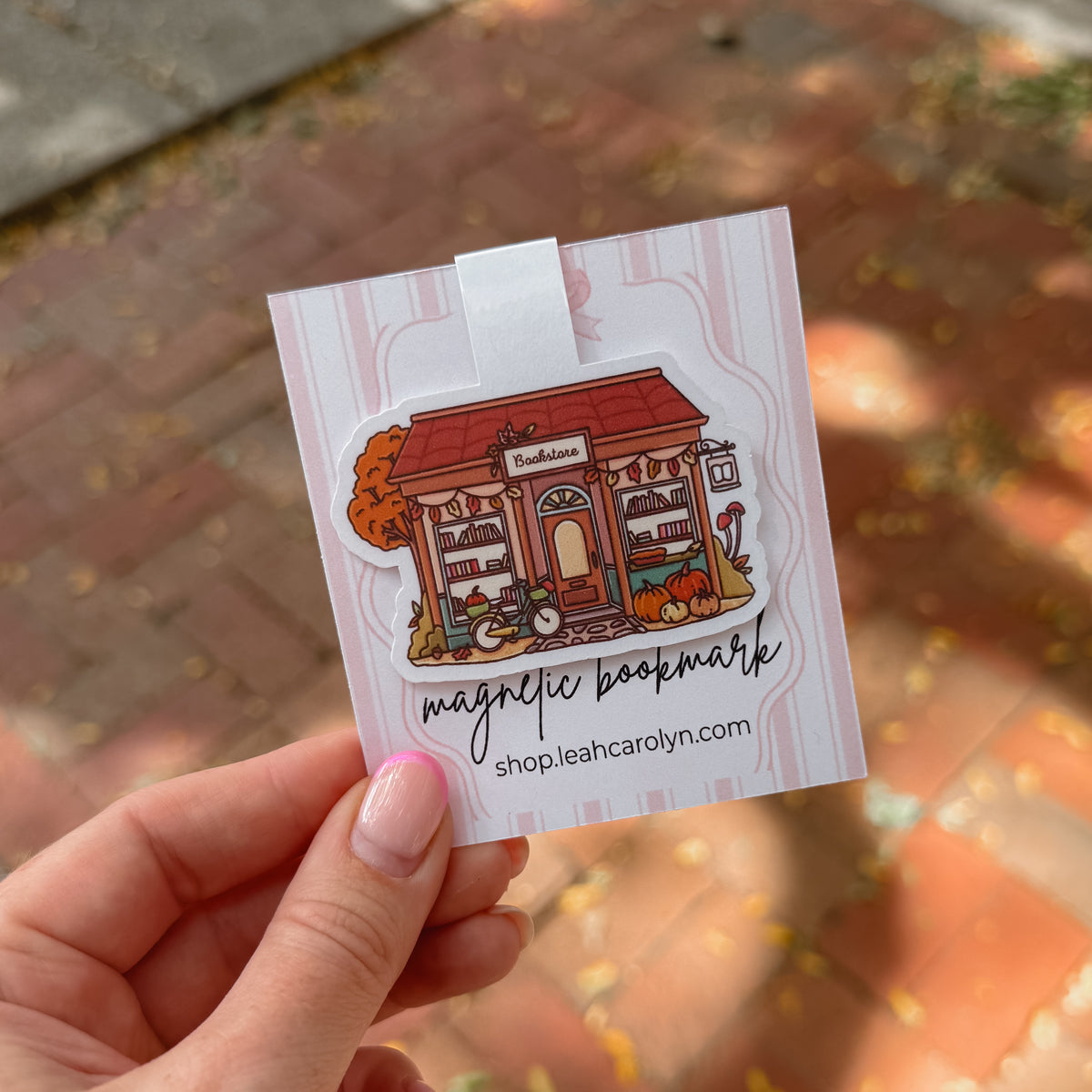 A hand with pink manicured nails holds a die-cut magnetic bookmark shaped like a storefront illustration. 