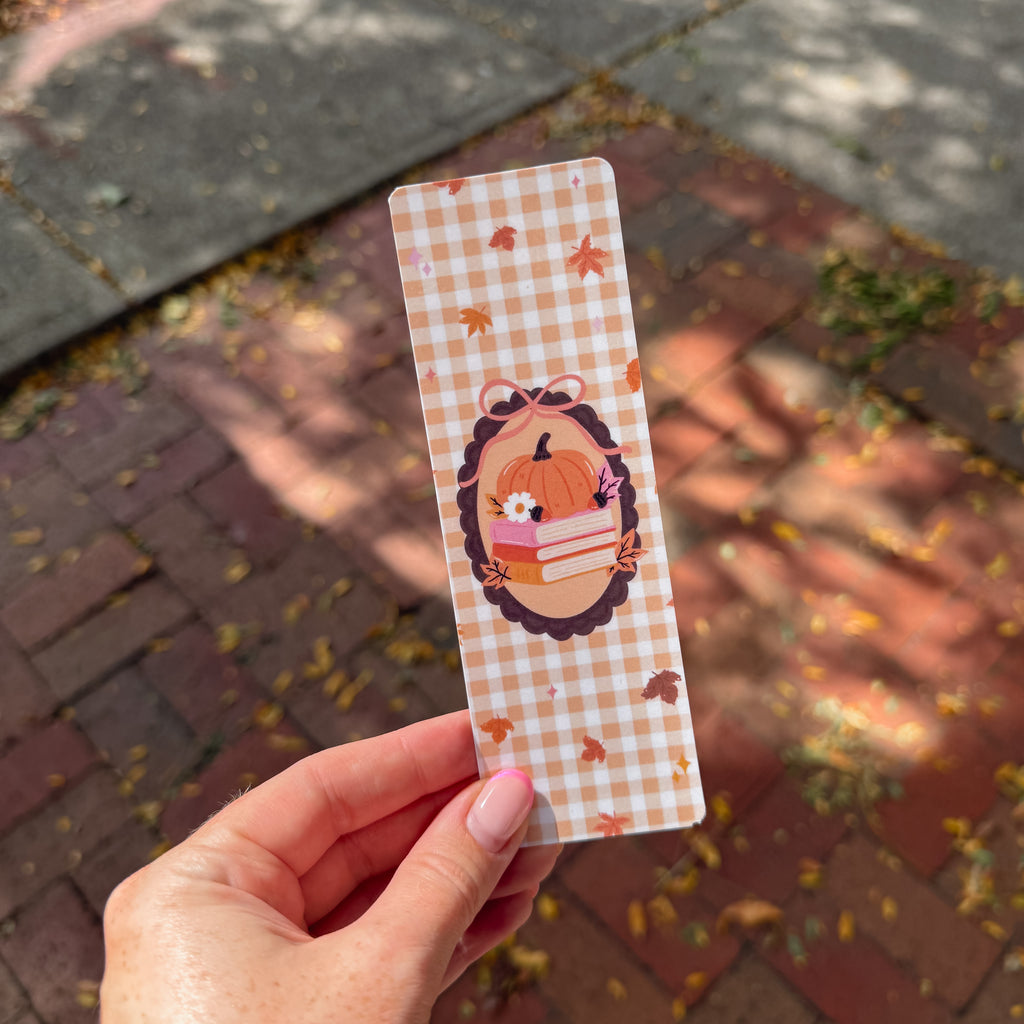 A hand with pink manicured nails holds a long, narrow autumn-themed bookmark featuring a white and orange gingham pattern, scattered fall leaves, and a central illustration of a pumpkin, books, and a flower.