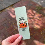 A hand with pink manicured nails holds a mint green bookmark outdoors on a brick path. The bookmark features a cute cartoon illustration of a ghost, pumpkin, and autumn leaves with the text 'Today's Forecast:'.