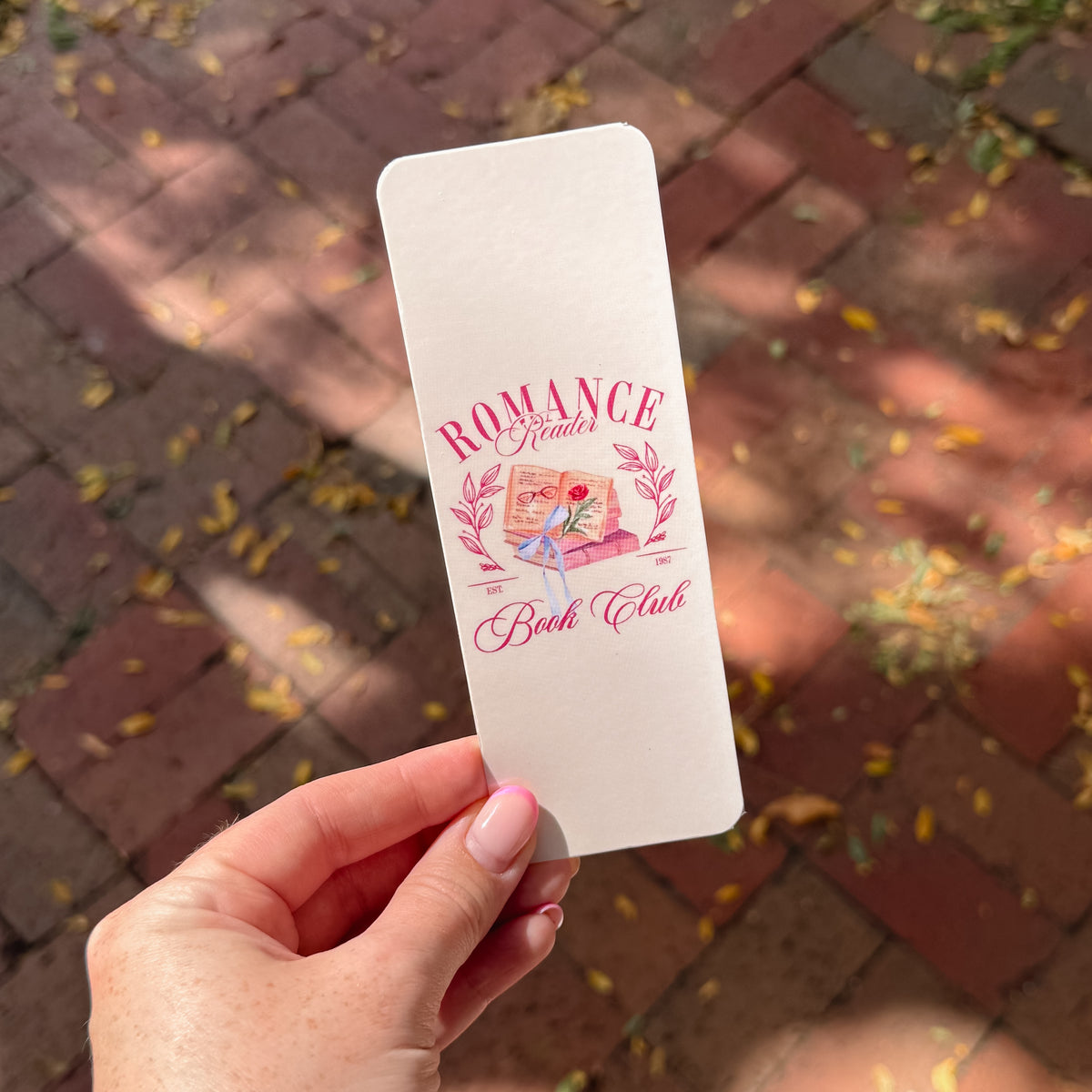 A hand holds a white rectangular bookmark featuring a pink and red illustration and the text 'ROMANCE Readers Book Club' against a blurred brick patio background with scattered leaves.