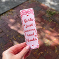 A hand holds a white paper bookmark outdoors over a brick pathway. The bookmark has red hand-drawn style text reading 'Girls just wanna read books' with small heart accents and a bow illustration at the top.