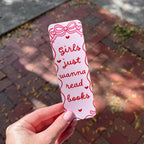 A hand holds a white paper bookmark outdoors over a brick pathway. The bookmark has red hand-drawn style text reading 'Girls just wanna read books' with small heart accents and a bow illustration at the top.