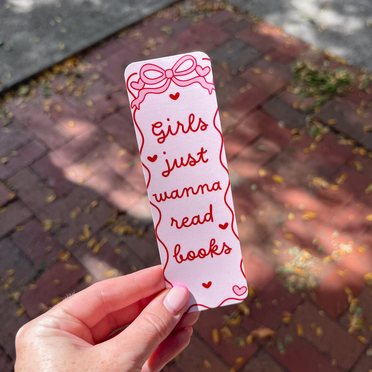 A hand holds a white paper bookmark outdoors over a brick pathway. The bookmark has red hand-drawn style text reading 'Girls just wanna read books' with small heart accents and a bow illustration at the top.