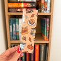 A hand with red nail polish holds a tan paper bookmark featuring a fall-themed illustration of a pumpkin, pancakes, autumn leaves, and a coffee sign in front of a wooden bookshelf.
