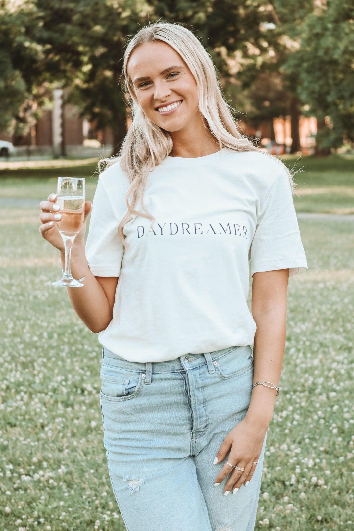 a photo of a girl in a park wearing a white t-shirt with daydreamer written in a modern deco font. She is smiling and holding a glass of champagne