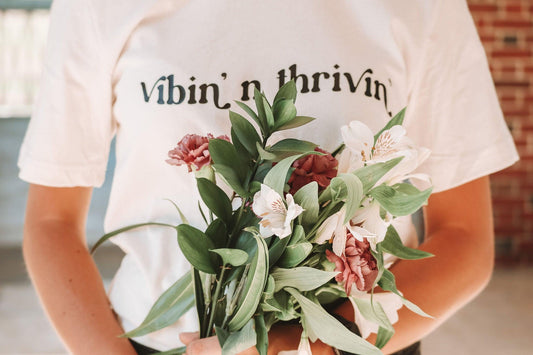 A close up photo of white and purple flowers and behind them a girl wearing a white short sleeve shirt with "vibin' n thivin'" printed in black wavy text across the front.