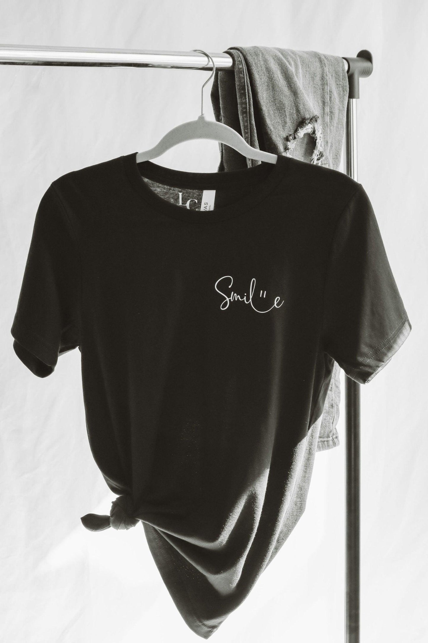 A black and white photo of a short sleeve T-shirt hanging on a clothing rack with a styled pair of hark jeans next to it. Smile is written in a cute cursive font, with a smiley face illustrated between the connection of l and e.