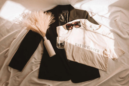 a flat lay photo of a white t-shirt with daydreamer written in a modern deco font. It is laying on top of a black blazer. There is a pair of sunglasses, a glass and a vase of dried pampas grass laying in the photo for decor