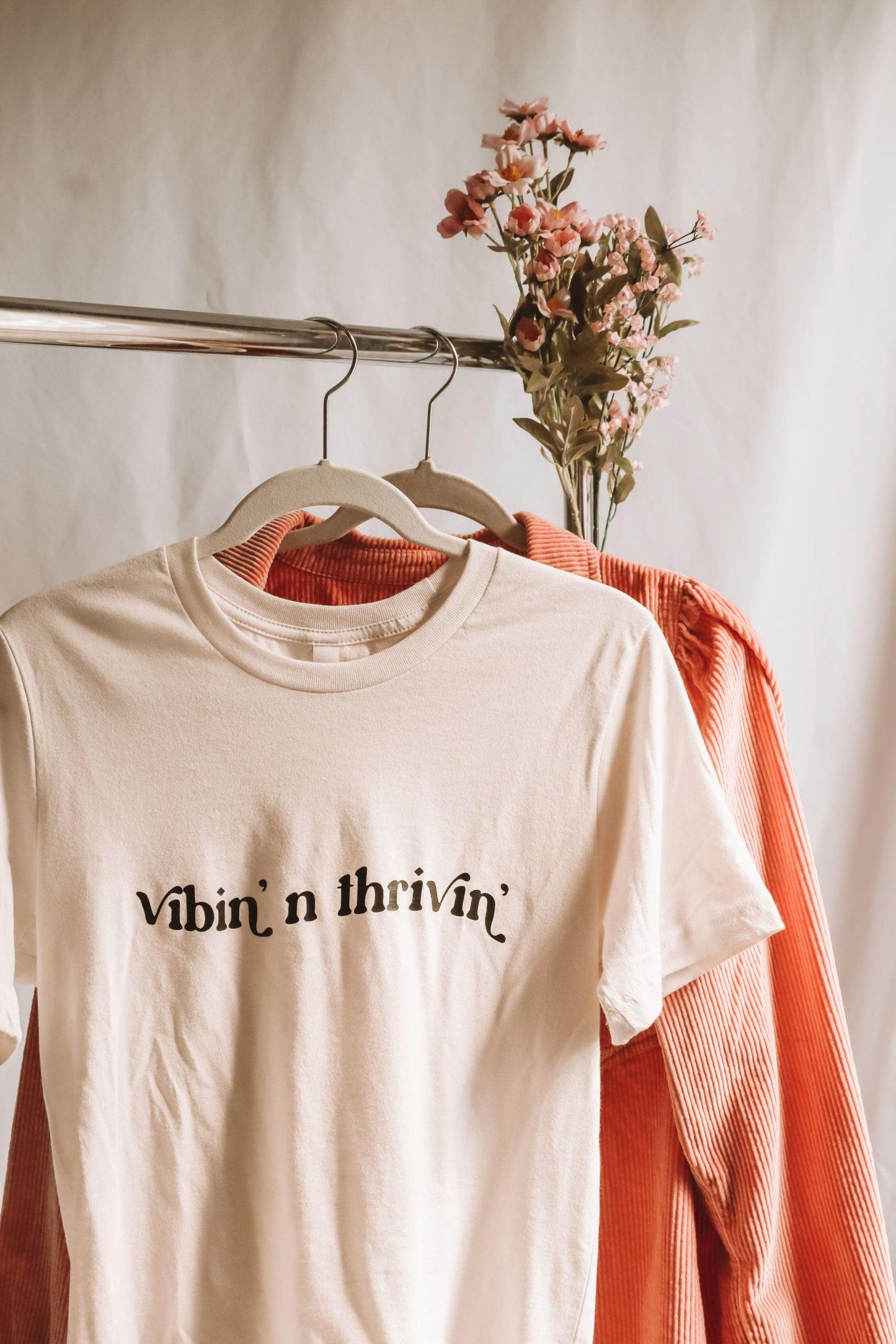 A white shirt with "vibin' n thrivin'" written in wavy retro text hanging on a clothing rack in front of a pink corduroy button up shirt