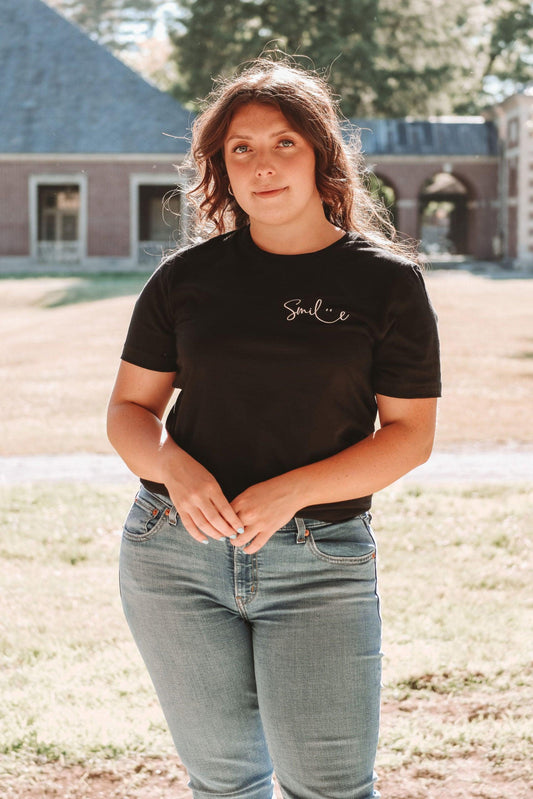 A girl with her hair pulled back wearing a black short sleeve T-shirt and light wash denim jeans outside in a park. Smile is written in a cute cursive font in the top left corner of the shirt where a pocket would be.