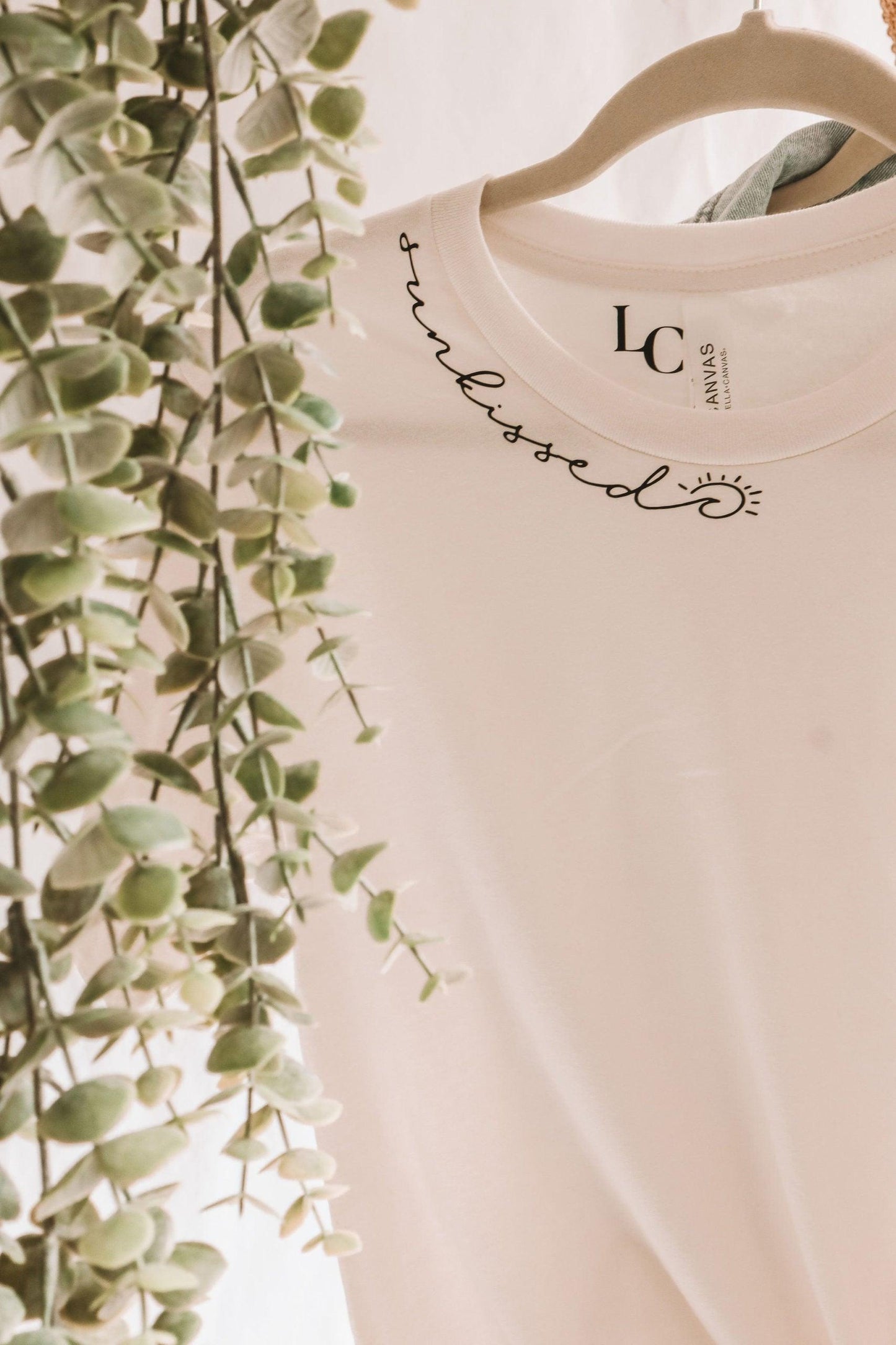 A close up photo of a white short sleeve t-shirt with sunkissed written in script text along the neckline. there's a wave and a sun in the script. It's hanging on a clothing rack with vines.