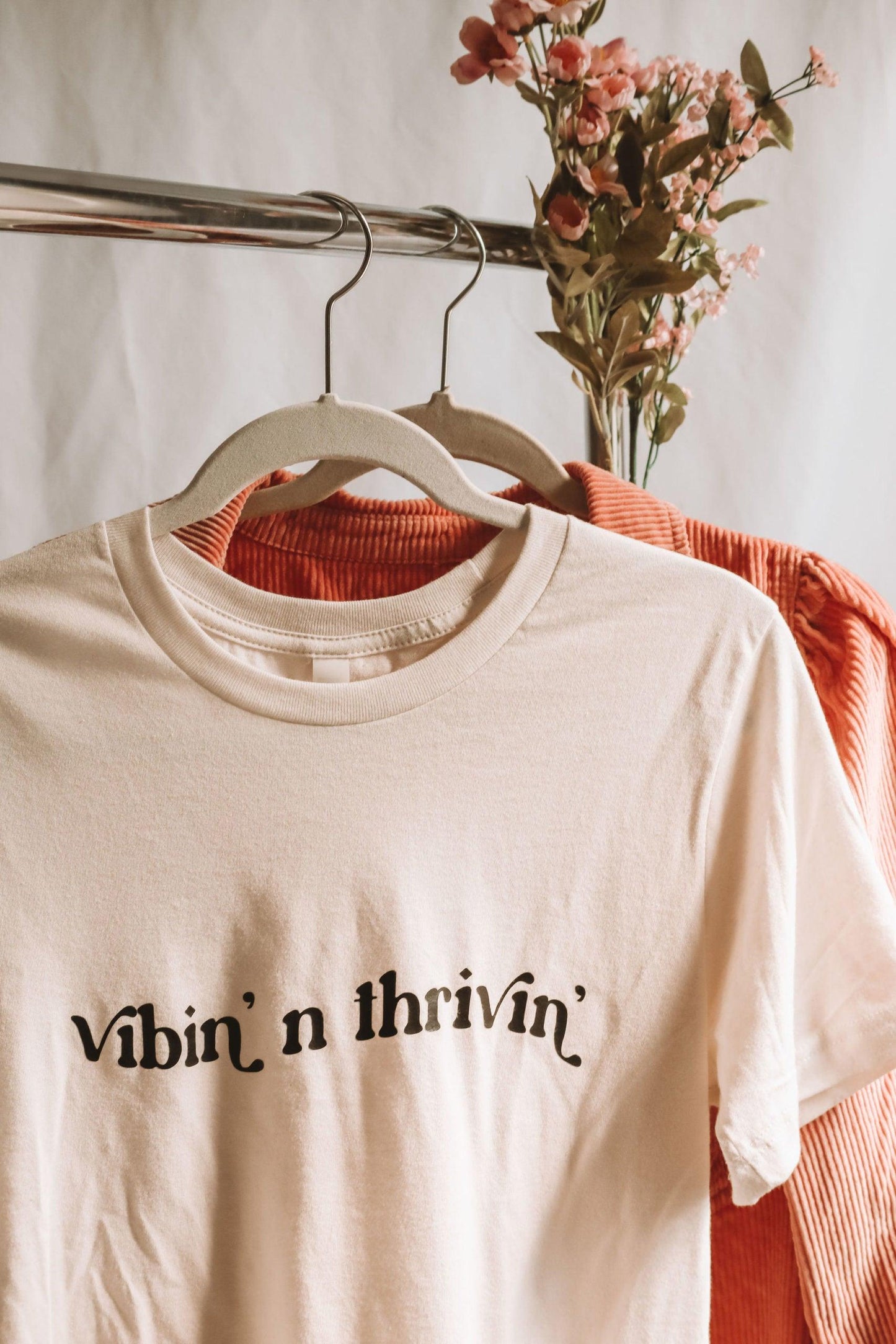 a closer cropped photo of a white shirt with "vibin' n thrivin'" written in wavy retro text hanging on a clothing rack in front of a pink corduroy button up shirt