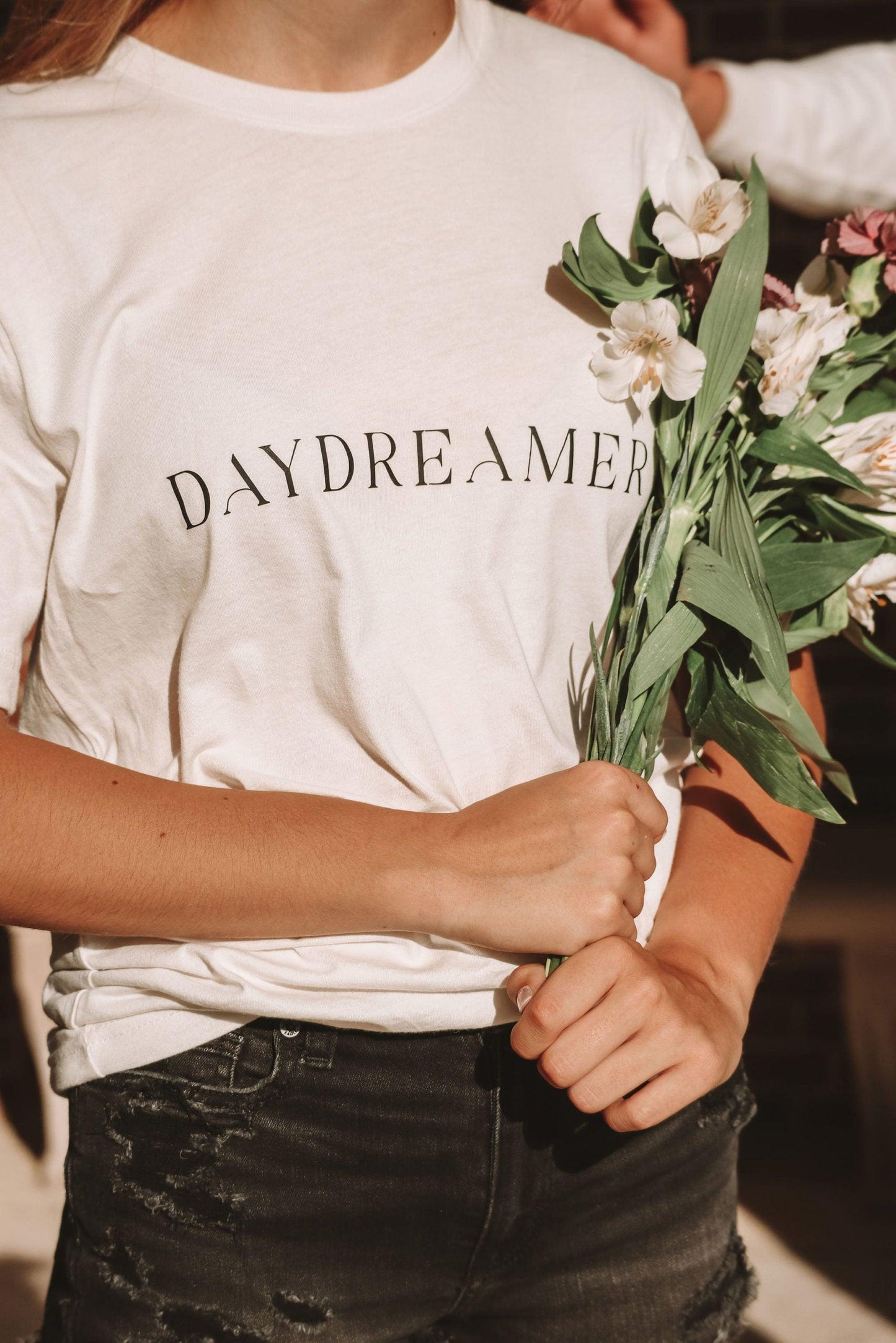 a cropped photo of a girl wearing a white t-shirt with daydreamer written in a modern deco font. She has white and purple flowers in her hands