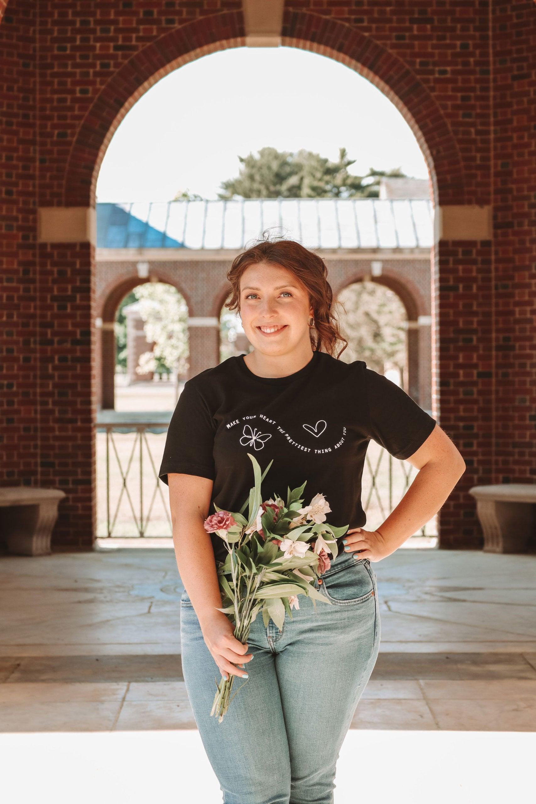 A photo of a girl in a black t-shirt and jeans. The shirt says "Make your heart the prettiest thing about you" with a butterfly and a heart. The girls has one hand on her hip and purple and white flowers in the other hand