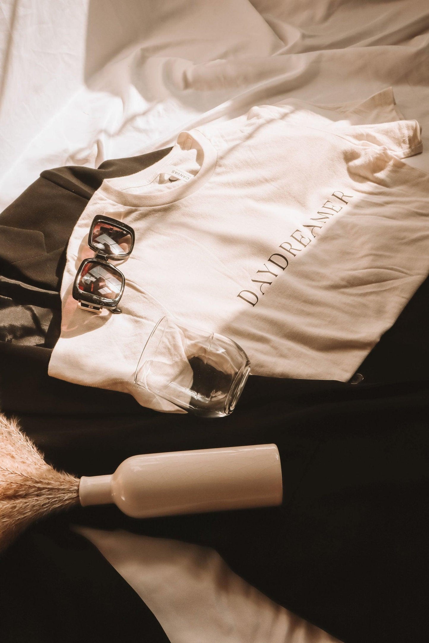 an angled photo of a white t-shirt with daydreamer written in a modern deco font. It is laying on top of a black blazer. There is a pair of sunglasses, a glass and a vase of dried pampas grass laying in the photo for decor