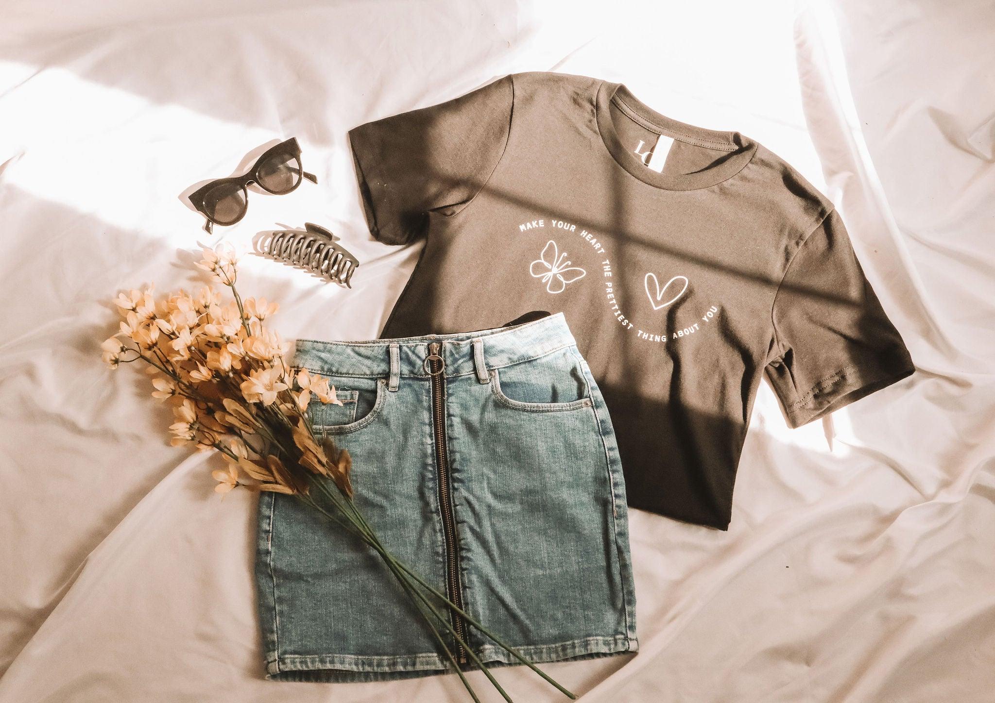 A flat lay photo of a black t-shirt and a jean skirt. The shirt says "Make your heart the prettiest thing about you" with a butterfly and a heart. There is a black claw clip, sunglasses and orange flowers as accessories