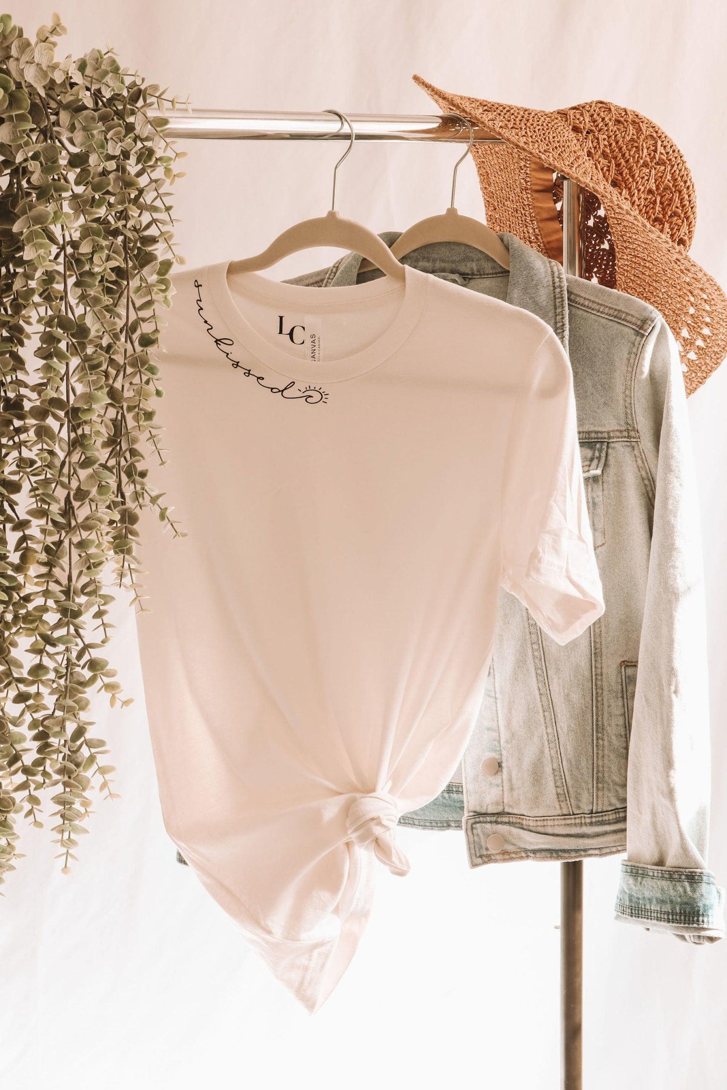 A white short sleeve t-shirt with sunkissed written in script text along the neckline. there's a wave and a sun in the script. The shirt is knotted at the bottom for styling. It's hanging on a clothing rack with a jean jacket, straw hat and vines.