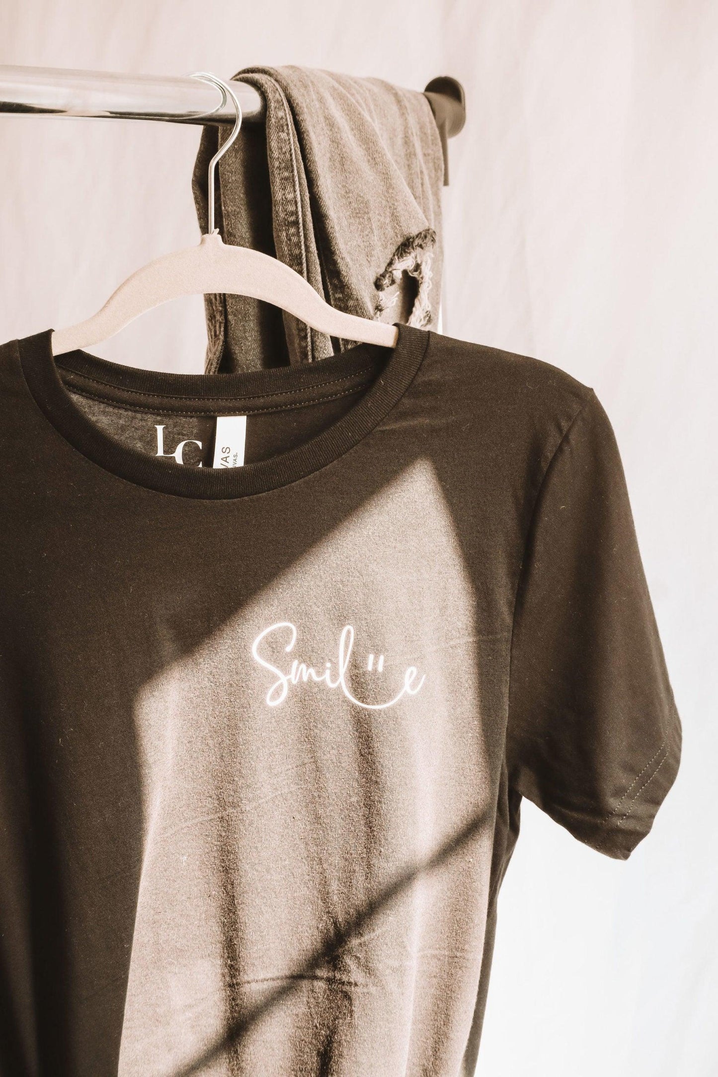 A black short sleeve T-shirt hanging on a clothing rack. Smile is written in a cute cursive font, with a smiley face illustrated between the connection of l and e. There is natural sunlight illuminating the design.