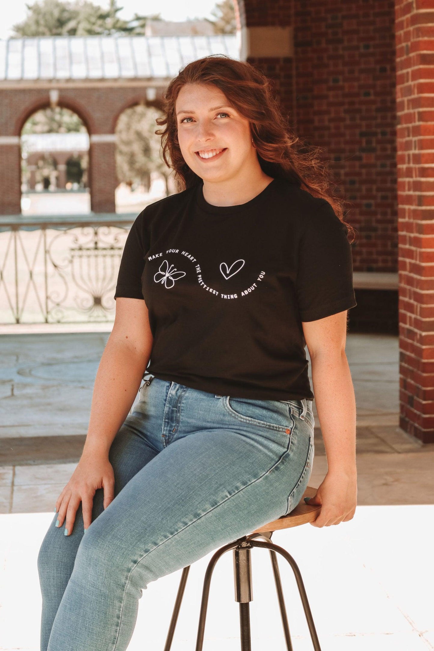 A photo of a girl in a black t-shirt and jeans. The shirt says "Make your heart the prettiest thing about you" with a butterfly and a heart. The girls is sitting on a stool at a slight angle