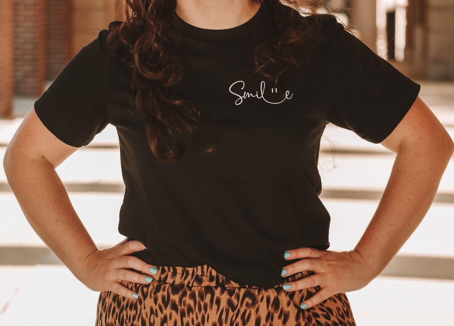 A cropped photos of a girl in the black smile t-shirt and a leopard skirt with her hands on her hips. The cursive text is in the upper left corner of the shirt where a pocket would be.