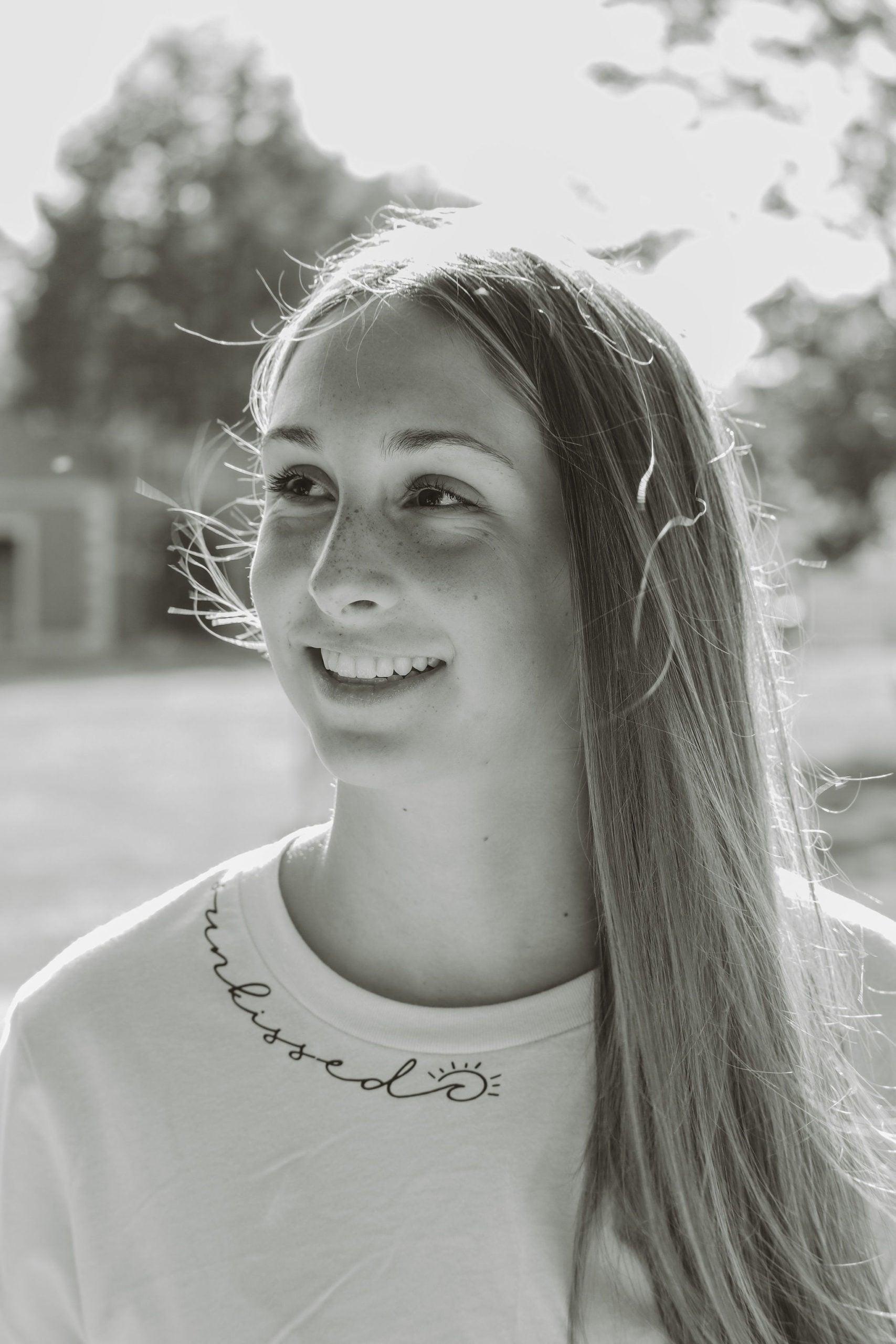 A black and white photo of a smiling girl in a park wearing a white short sleeve t-shirt with sunkissed written in script text along the neckline. there's a wave and a sun in the script.