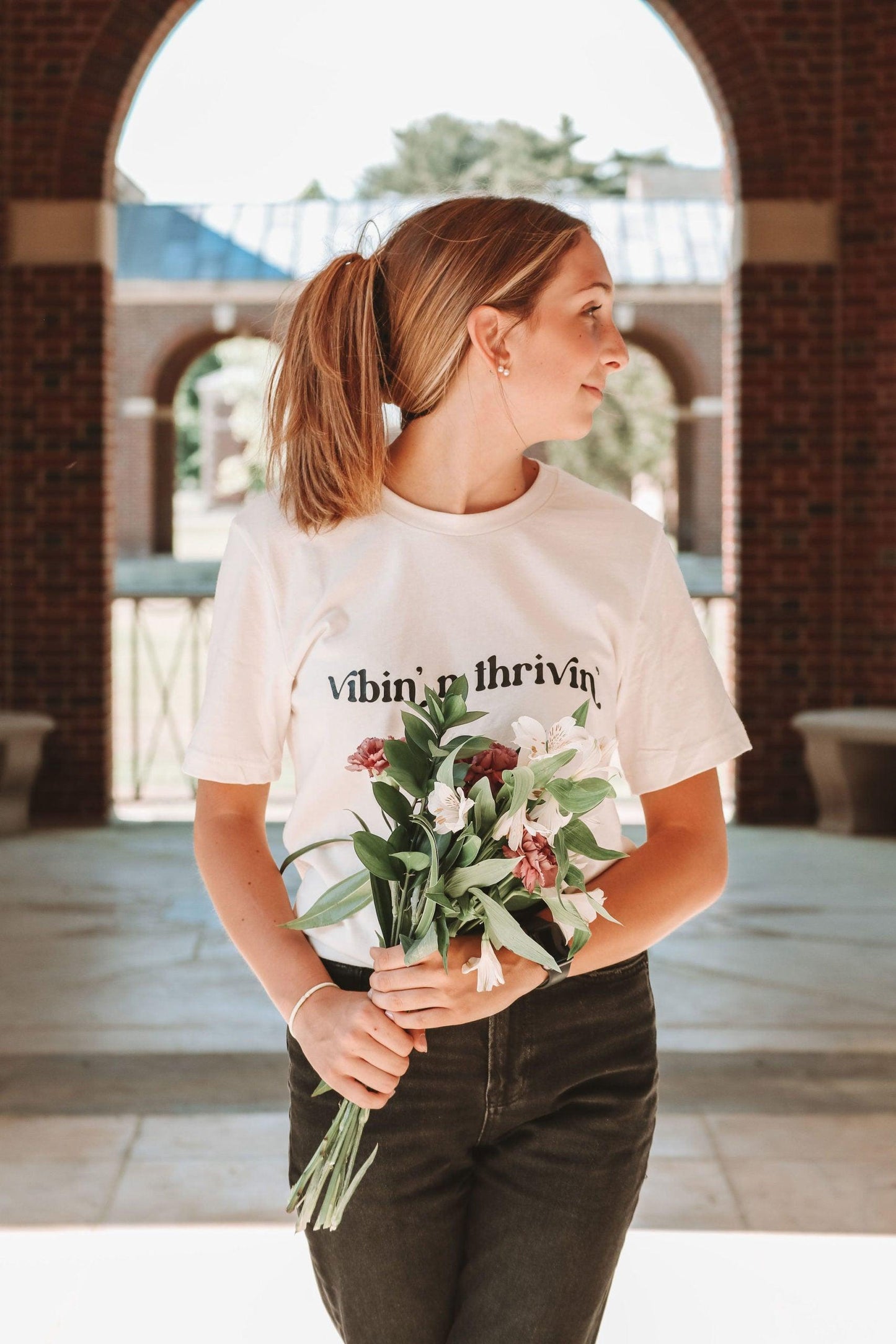 A photo of a girl with her hair clipped up, holding white and purple flowers. She is wearing a white short sleeve shirt with "vibin' n thivin'" printed in black wavy text across the front.