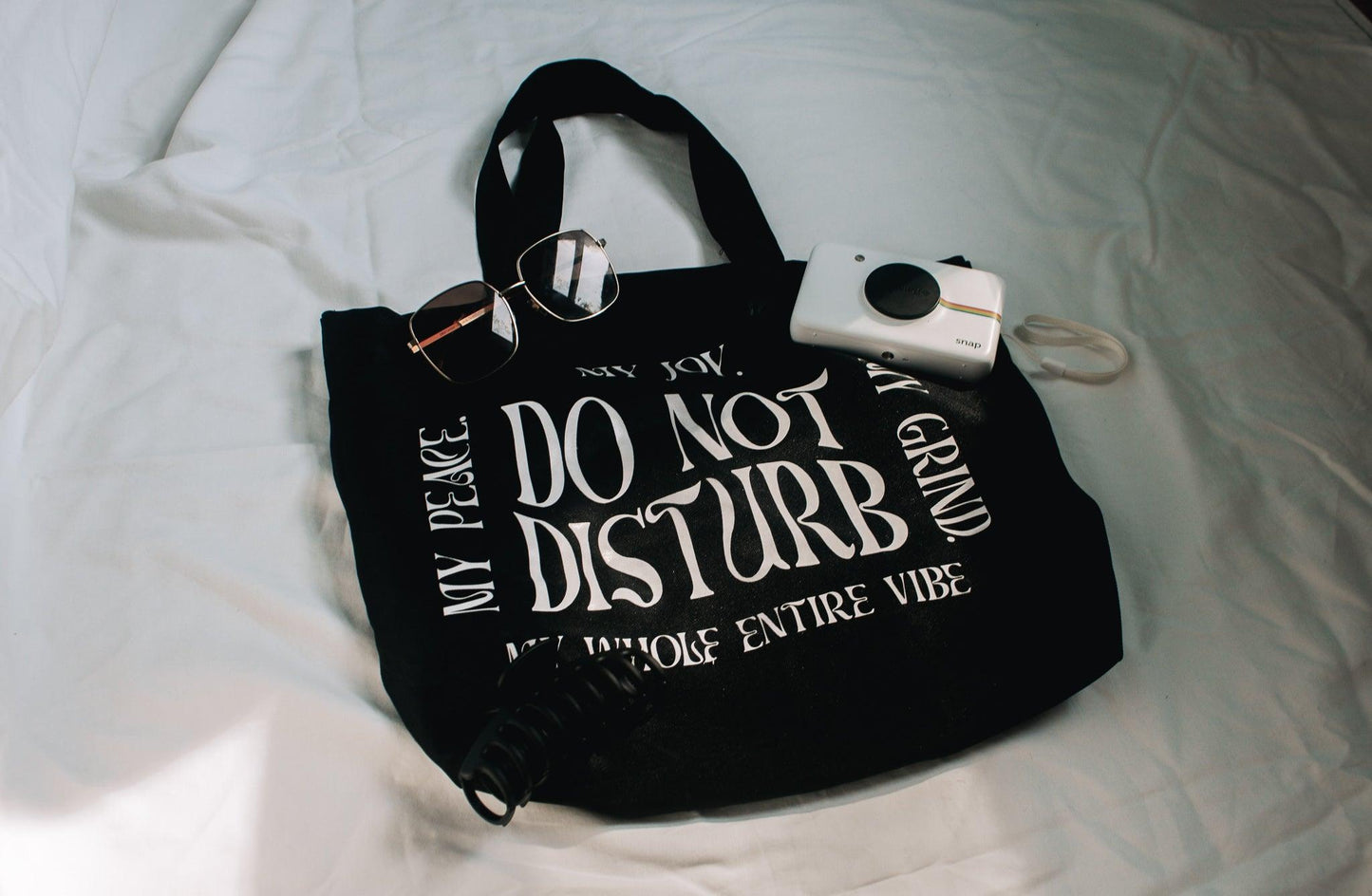 An angled photo of the do not disturb tote bag against a white background with a polaroid, sunglasses and a claw hair clip laying on and around it for styling.
