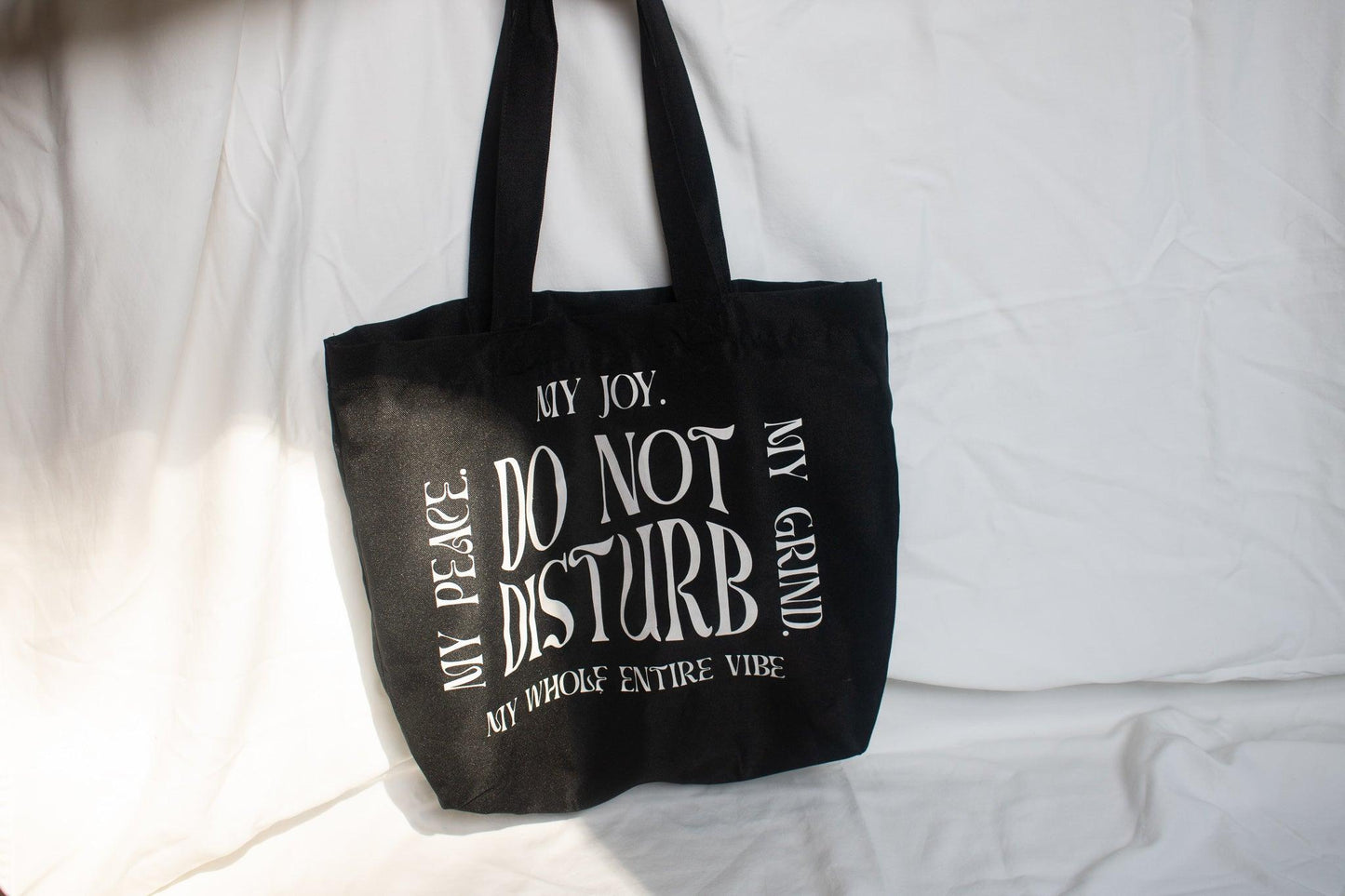 A photo of a hanging black tote bag against a white background that has do not disturb in the middle. Along the sides going clockwise forming a square is "My Joy. My Grind. M whole entire vibe. My Peace." all in a retro wavy text.