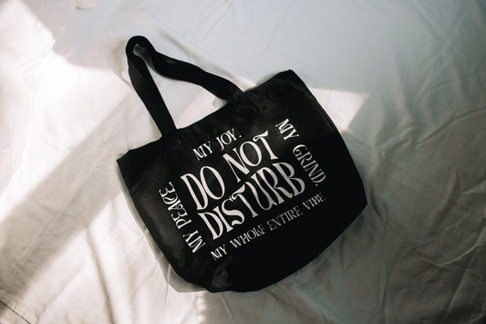 An angled photo of a black tote bad that has do not disturb in the middle. Along the sides going clockwise forming a square is "My Joy. My Grind. M whole entire vibe. My Peace." all in a retro wavy text.
