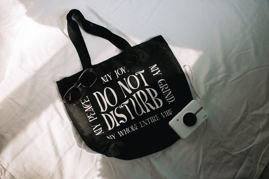 An angled photo of the do not disturb tote bag against a white background with a polaroid and sunglasses laying on and around it for styling.