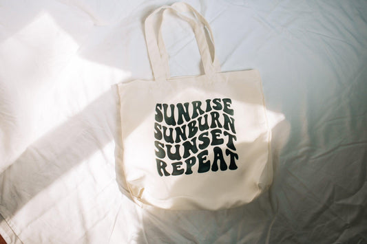 A beige tote bag is laying on a white sheet. There is natural light across the bag. The bag says sunrise, sunburn, sunset, repeat in groovy text.