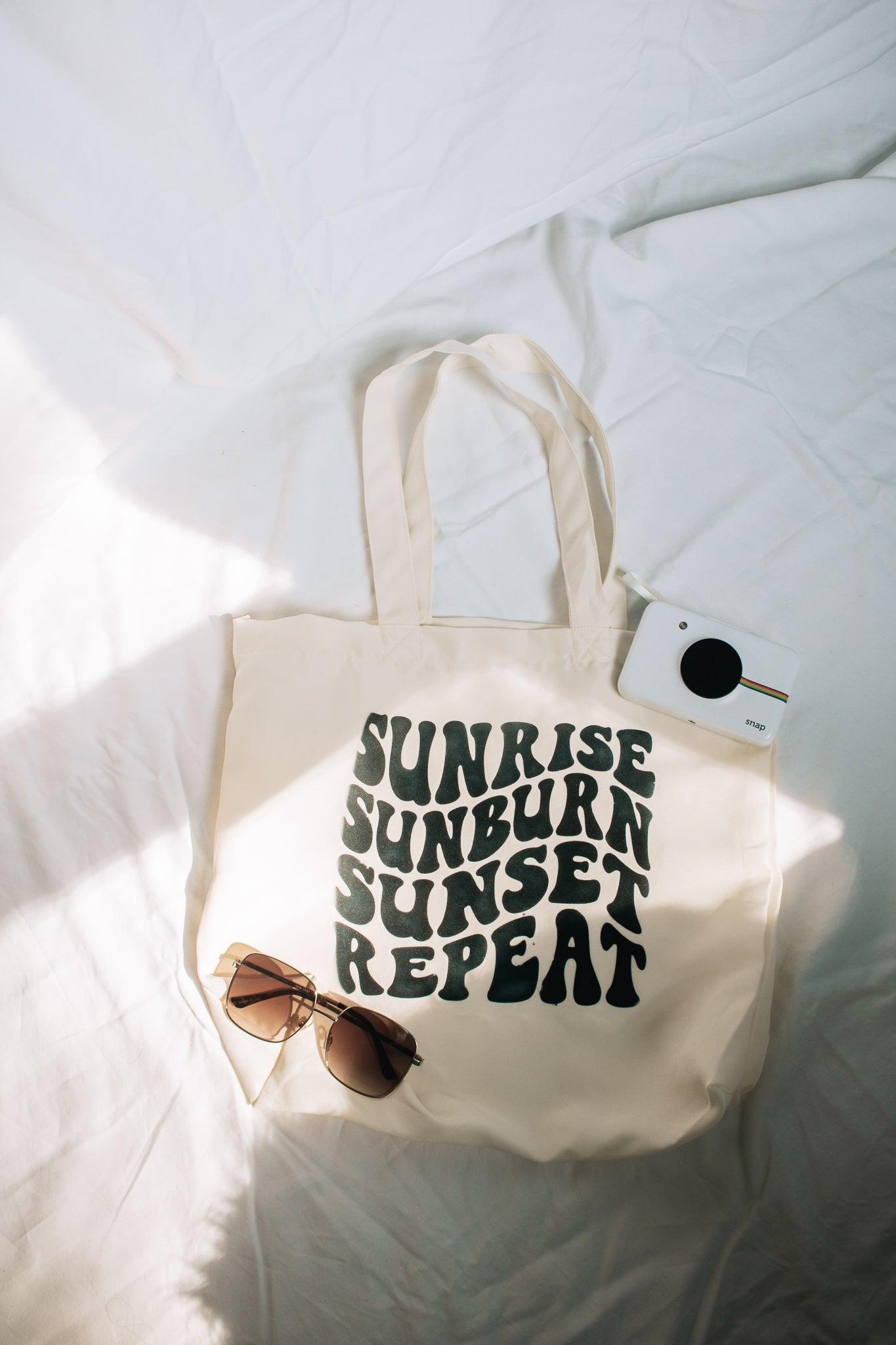 A portrait photo of a beige tote bag laying on a white sheet. The bag says sunrise, sunburn, sunset, repeat in groovy text. There is a polaroid camera and sunglasses laying on and around the bag for styling.