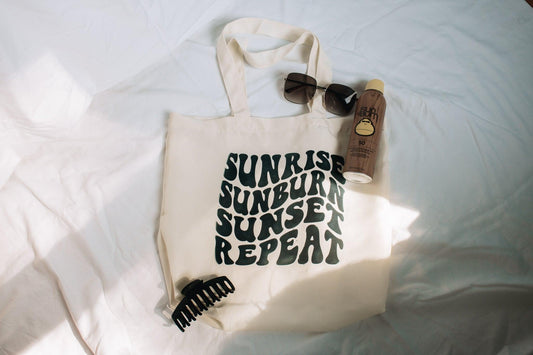 A portrait photo of a beige tote bag laying on a white sheet. The bag says sunrise, sunburn, sunset, repeat in groovy text. There is a claw hair clip, sunglasses, sunscreen laying on and around the bag for styling.