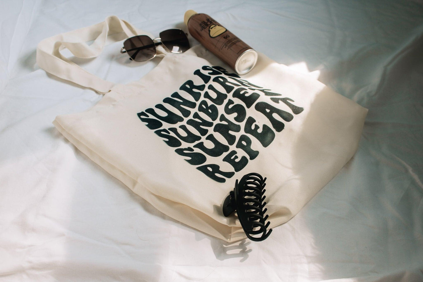 An angled photo of a beige tote bag laying on a white sheet. The bag says sunrise, sunburn, sunset, repeat in groovy text. There is a claw hair clip, sunglasses, sunscreen laying on and around the bag for styling.